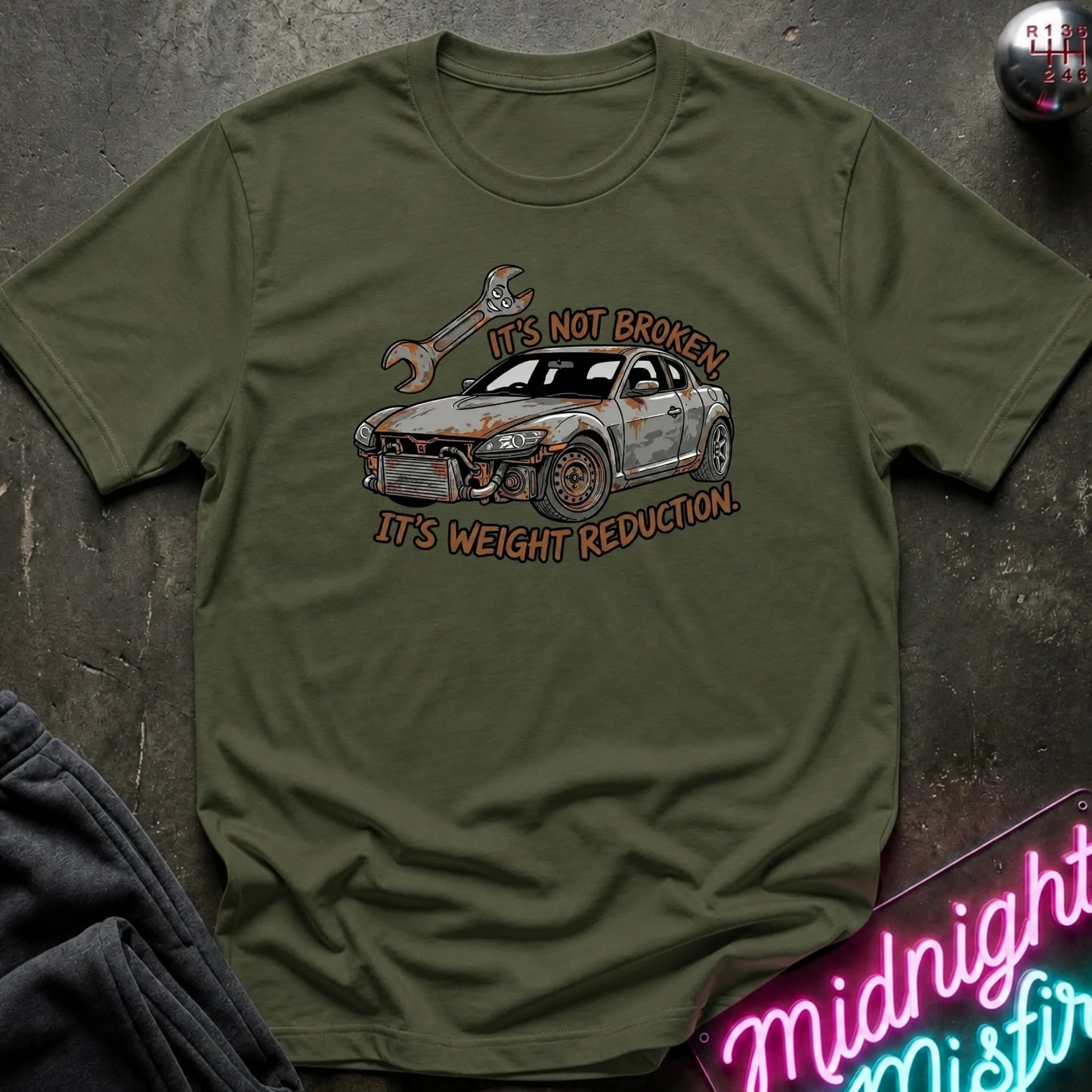 An olive green short-sleeve t-shirt laid flat on a dark textured surface. The shirt features a graphic of a rusted-out car with a wrench and the text 'IT'S NOT BROKEN.