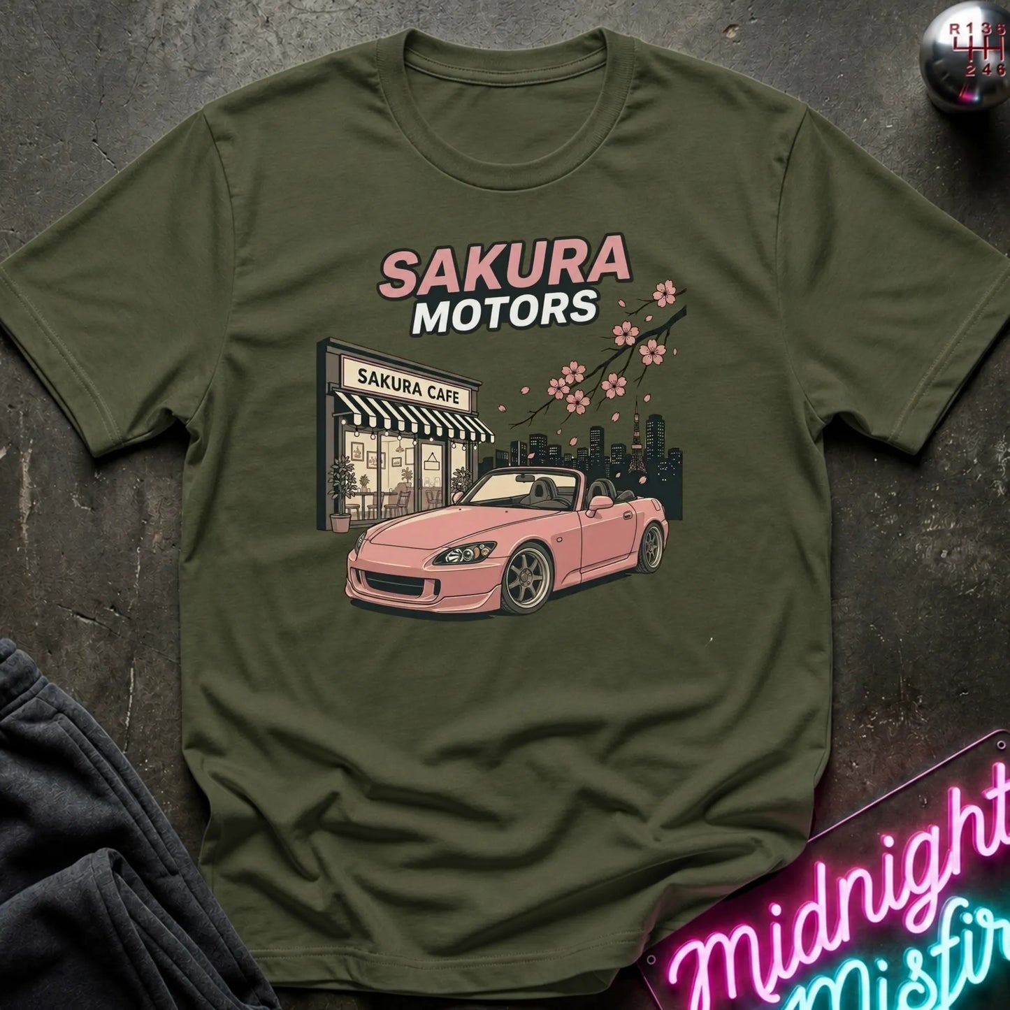 An olive green short-sleeve t-shirt laid flat on a dark textured surface, featuring a graphic print of a pink convertible car in front of a 'Sakura Motors' cafe with cherry blossoms.