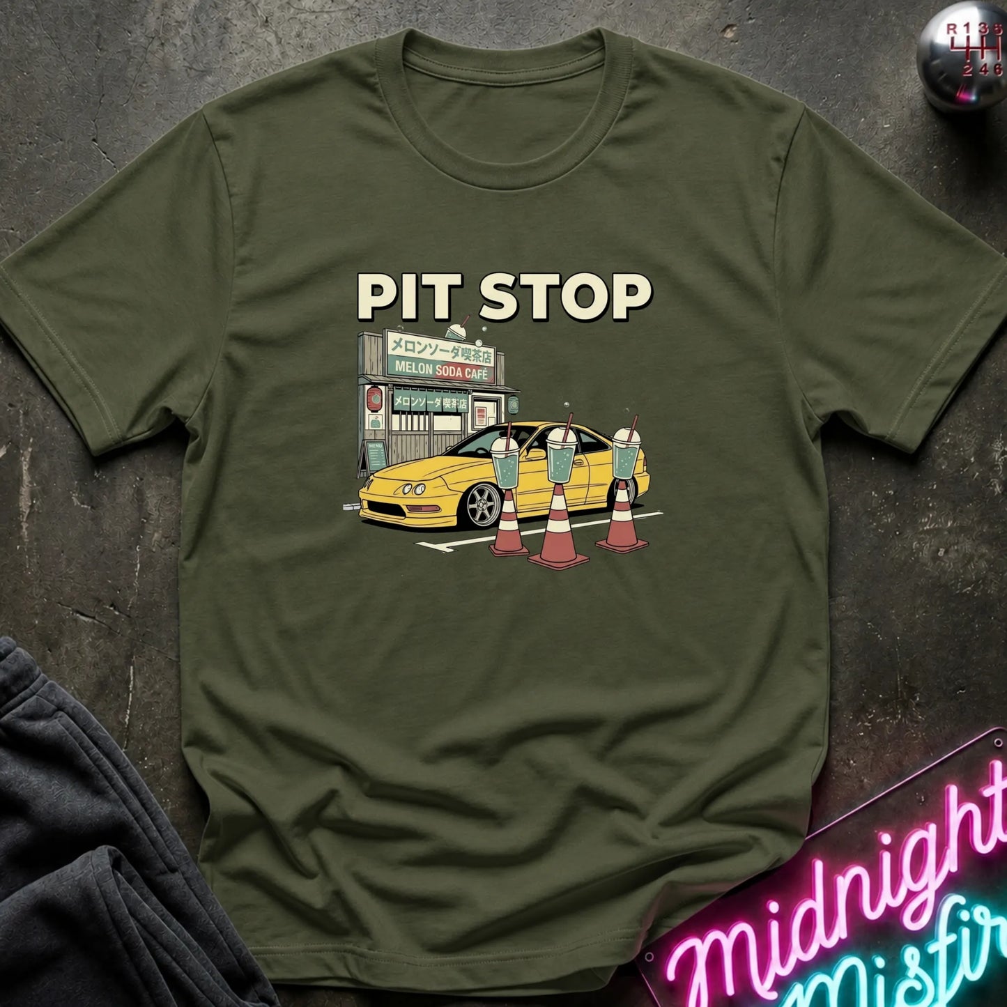 An olive green graphic t-shirt laid flat on a dark textured surface. The graphic features the text 'PIT STOP' above a cartoon illustration of a yellow car at a care with traffic cones.