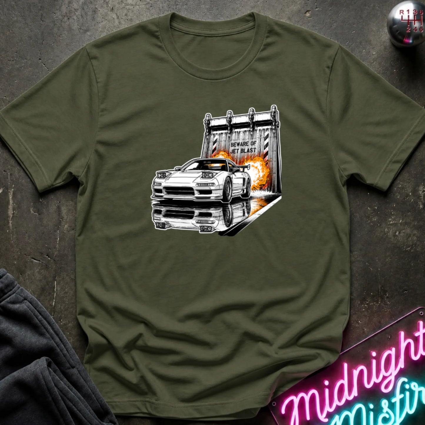A olive green short-sleeve t-shirt laid flat on a dark textured surface, featuring a graphic print of a white sports car in front of a blast off area.