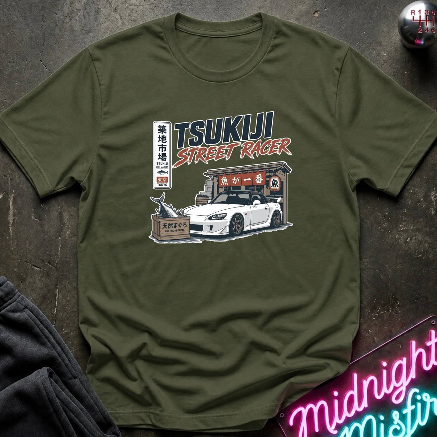 Olive green short-sleeve graphic T-shirt laid flat on and surrounding an illustration of a white sports car parked outside a fish market.