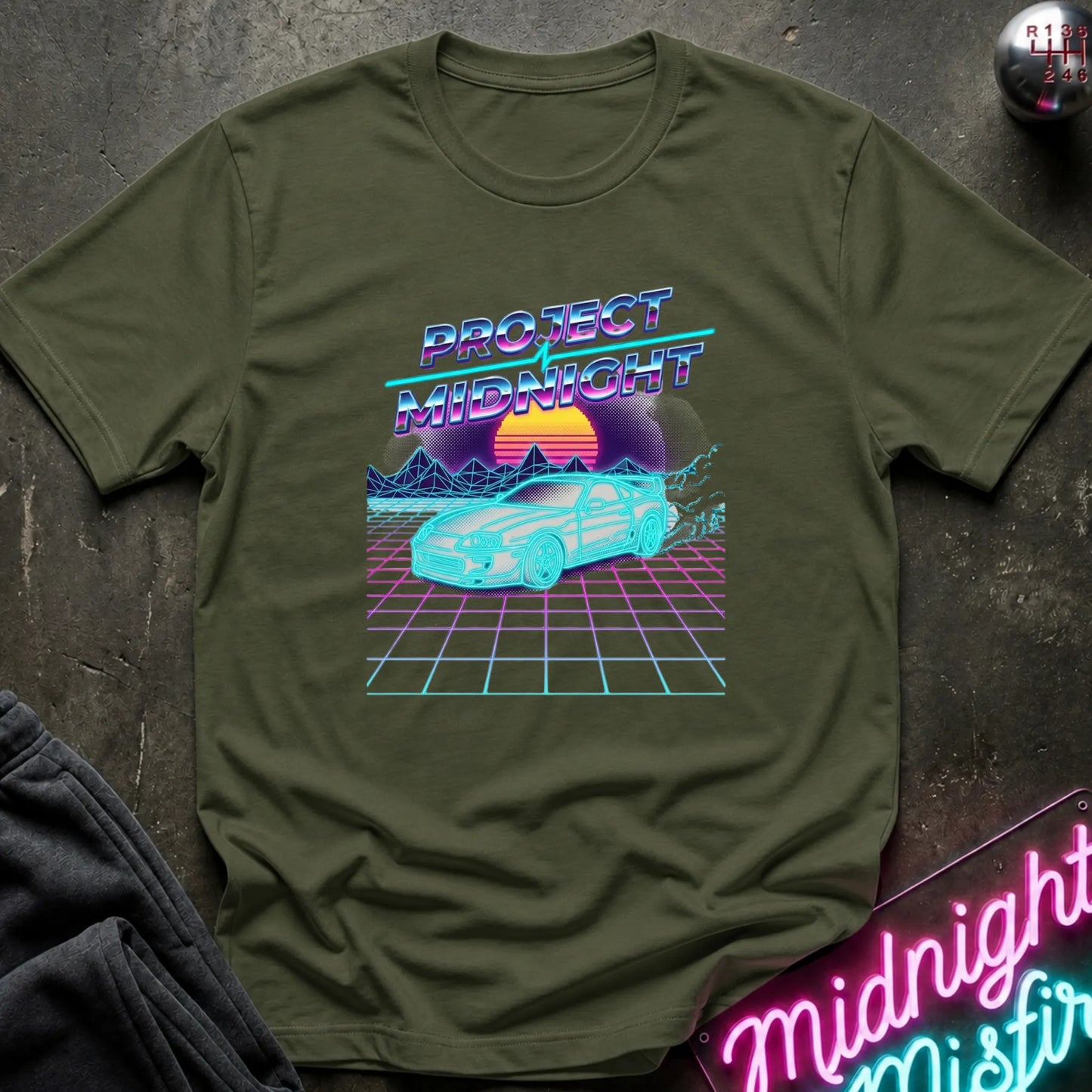 An olive green short-sleeve t-shirt with a retro 80s synthwave graphic reading 'PROTECT MIDNIGHT' above a cyan car and neon sunset, laid flat on a dark textured surface with a neon sign and dark pants partially visible.