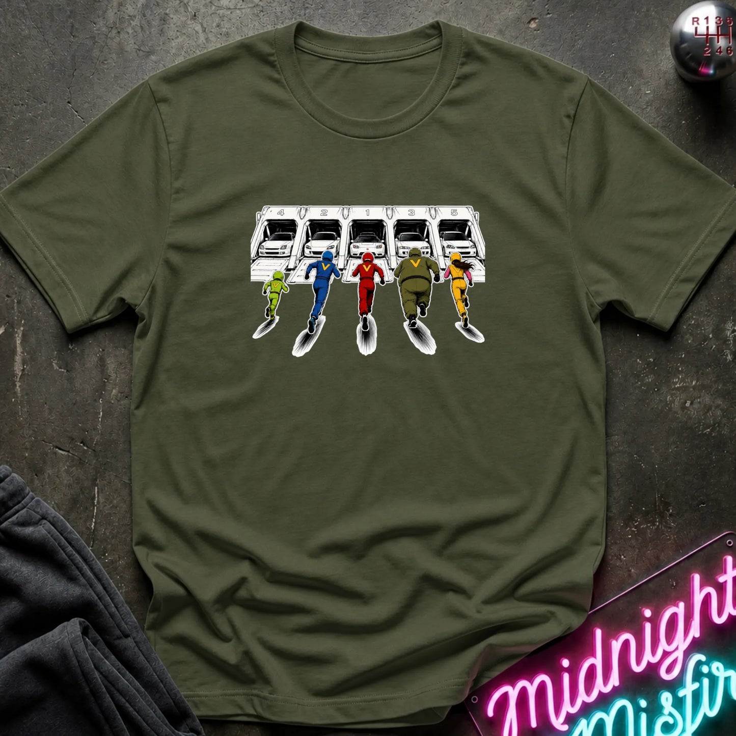An olive green short-sleeve graphic T-shirt laid flat on a dark textured surface. The shirt features a printed illustration of a white sports cars in 5 different launch pods.