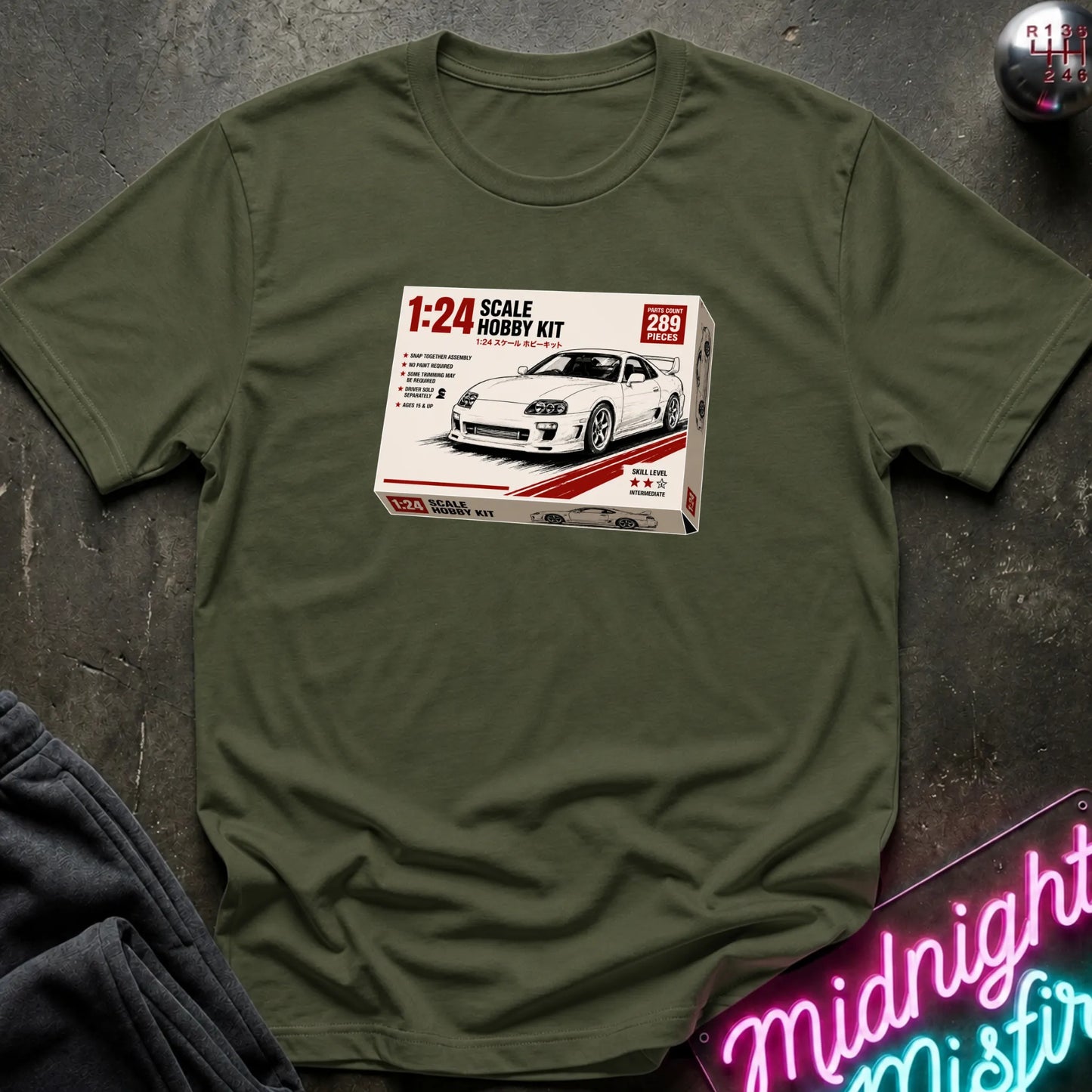 An olive green crewneck t-shirt laid flat on a dark textured surface, featuring a graphic print of a 1:24 scale model kit box for a white sports car.