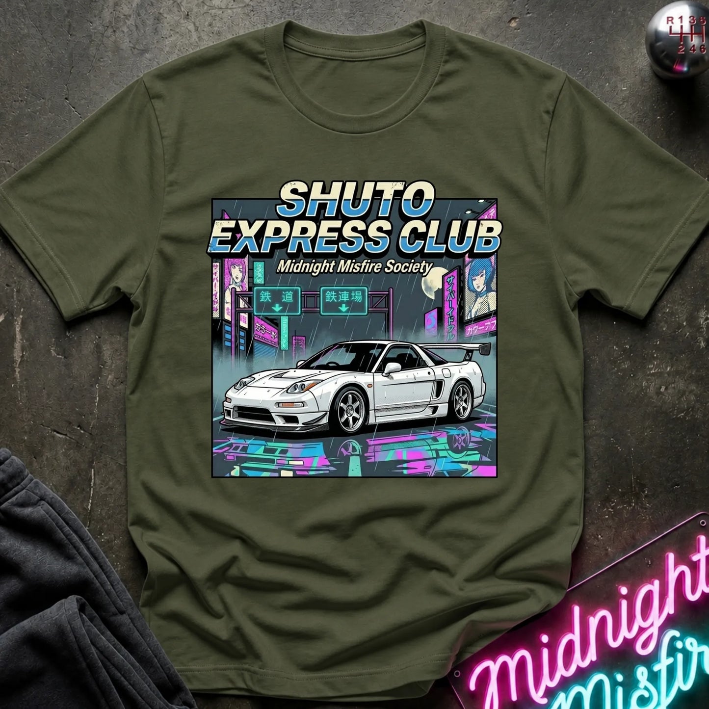 An olive green crewneck t-shirt featuring a colorful graphic print of a white sports car in a neon cyberpunk city with the text 'SHUTO EXPRESS CLUB' and 'Midnight Misfire Society'. The shirt is displayed flat on a dark textured surface with decorative props.