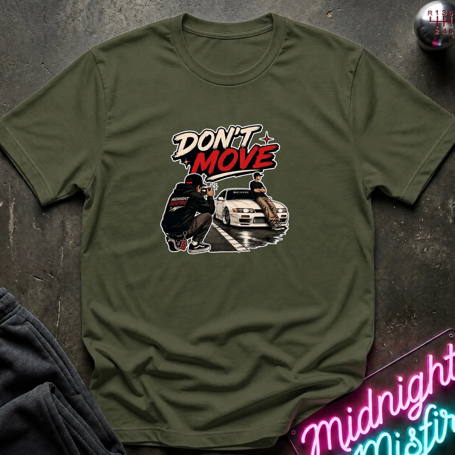 An olive green short-sleeve t-shirt with a graphic print on the front. The graphic features the text 'DON'T MOVE' in stylized white and red lettering above an illustration of a person crouching next to a white sports car.
