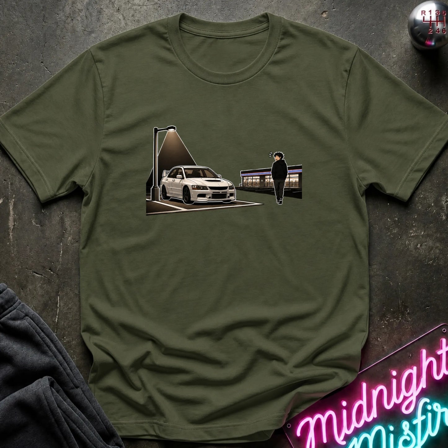 An olive green crewneck t-shirt laid flat on a dark textured surface, featuring a graphic print of a white sports car under a spotlight with a silhouette figure standing nearby. A neon sign and a pair of dark jeans are partially visible as props.