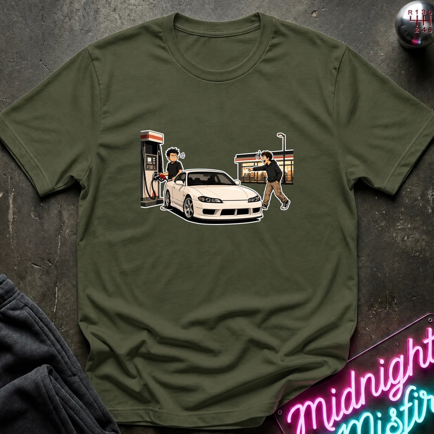 An olive green crewneck t-shirt featuring a graphic print of a white sports car at a gas station, styled flat-lay on a dark textured surface with a neon sign and dark sweatpants.