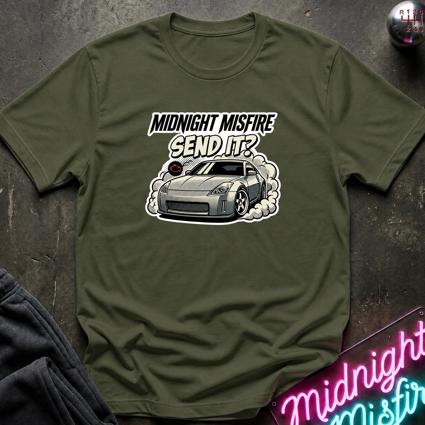 An olive green short-sleeve t-shirt laid flat on a dark textured surface, featuring a graphic of a silver sports car with smoke and the text 'MIDNIGHT MISFIRE SEND IT?'. A neon sign and dark fabric are partially visible in the corners.