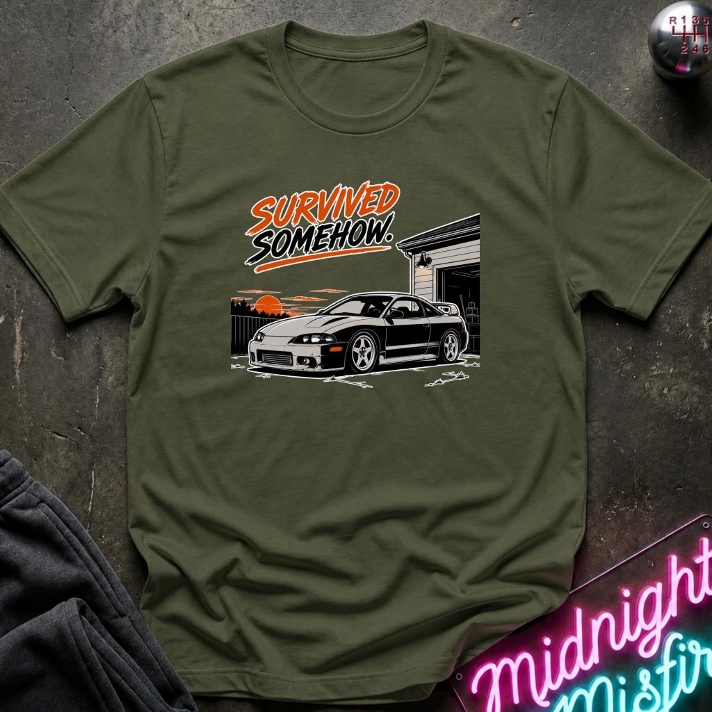 An olive green crewneck t-shirt laid flat on a dark textured surface, featuring a graphic print of a black sports car in front of a garage with the text 'SURVIVED SOMEHOW.' in orange and white.