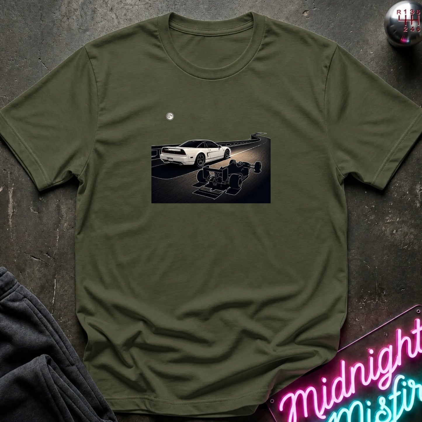 An olive green short-sleeve crewneck t-shirt laid flat on a dark textured surface, featuring a graphic print of a white sports car and a shadow car on a night road scene. The shirt is styled with dark gray sweatpants, a silver ball, and a neon sign in the corners.