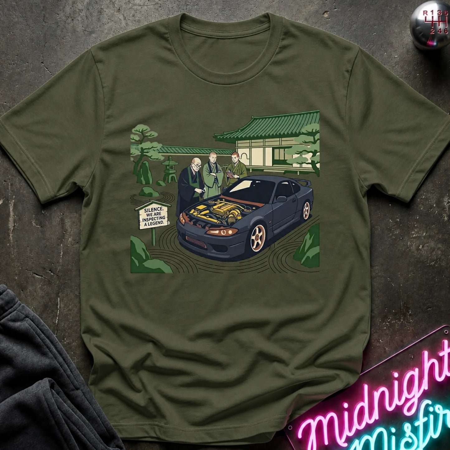 Olive green short-sleeve graphic t-shirt laid flat on a dark textured surface. The graphic shows a black sports car with its hood open, surrounded by two men in suits, a Japanese-style building, and green trees.