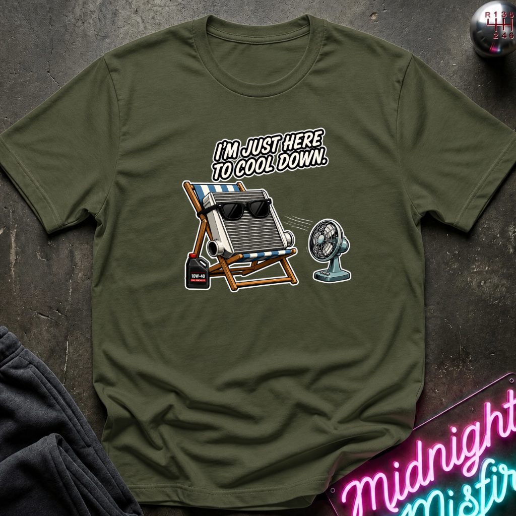 An olive green graphic t-shirt laid flat on a dark textured surface. The graphic features a retro beach chair with a radiator wearing sunglasses, a small fan, and the text 'I'M JUST HERE TO COOL DOWN.