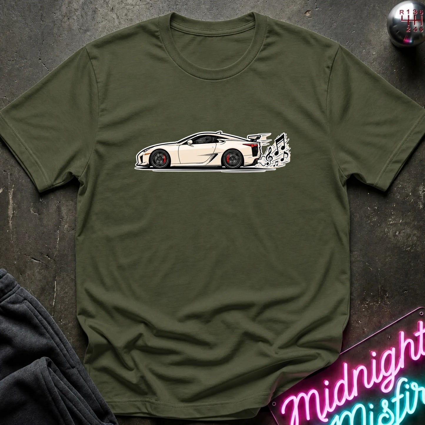 An olive green short-sleeve t-shirt laid flat on a dark textured surface, featuring a graphic print of a white sports car with a music note coming from exhaust.
