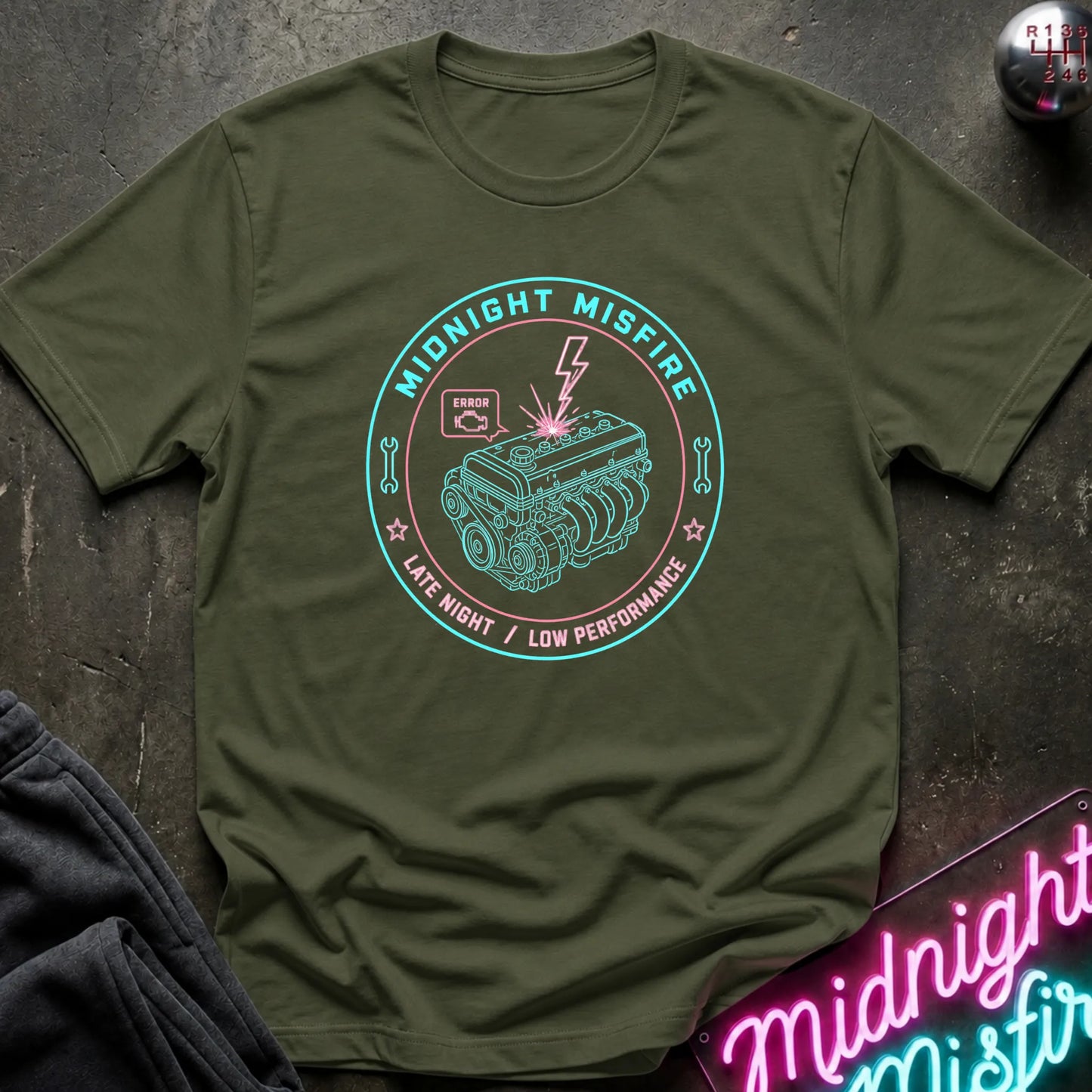 An olive green short-sleeve t-shirt with a circular graphic on the chest reading 'Midnight Misfire' and showing an engine illustration with a lightning bolt. The shirt is laid flat on a dark textured surface with a neon sign and dark pants partially visible as props.