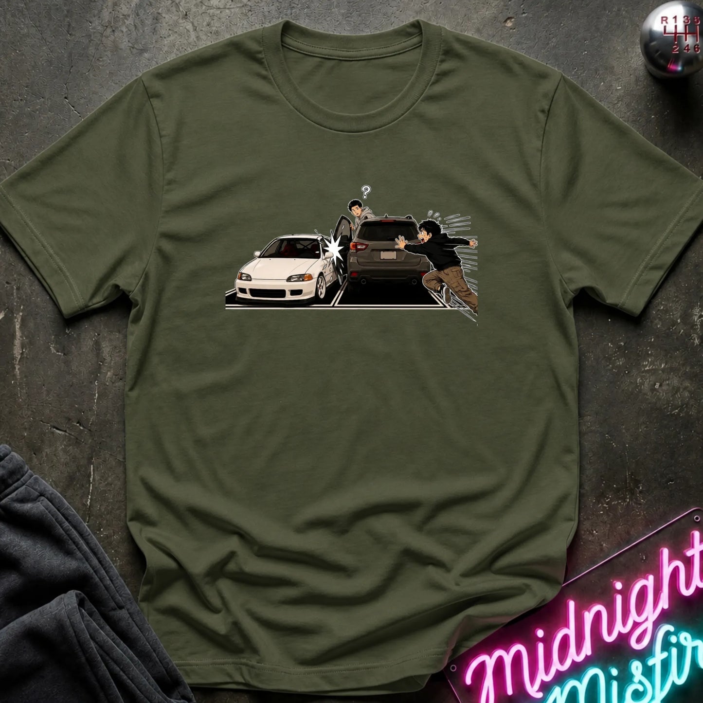 An olive green short-sleeve t-shirt laid flat on a dark textured surface, featuring a graphic print of a white sports car and a black SUV with two illustrated characters in the center. A neon sign and folded dark fabric are partially visible around it.