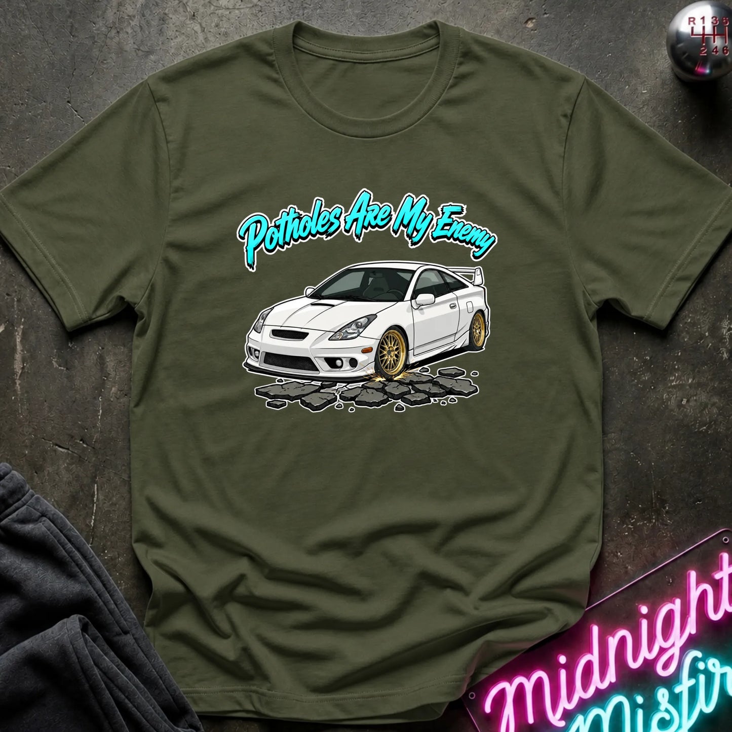 An olive green short-sleeve graphic T-shirt laid flat on a dark textured surface. The shirt features a white car illustration with gold wheels and the phrase 'Potatoes Are My Enemy' in teal script above it.