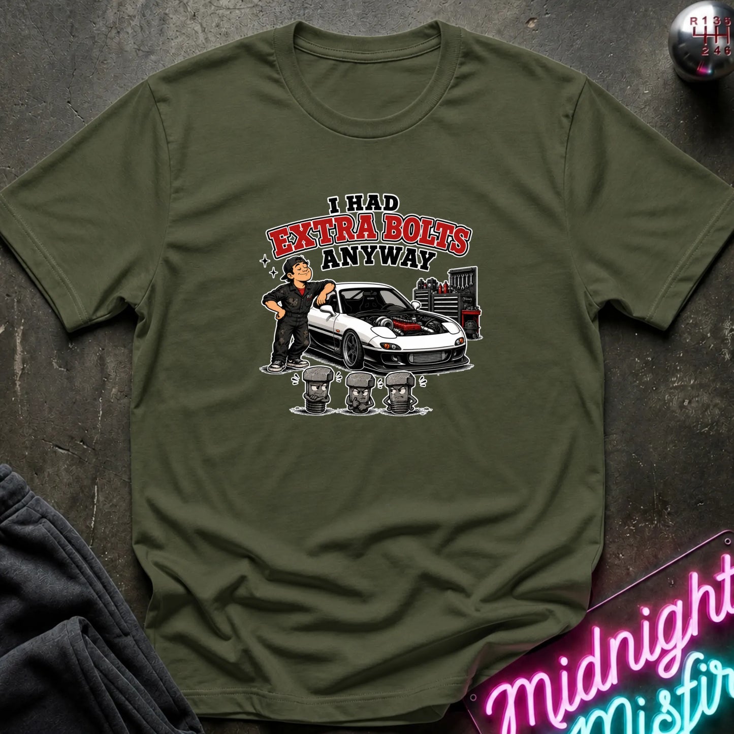 An olive green short-sleeve graphic t-shirt laid flat on a dark textured surface. The graphic features a cartoon of a man standing next to a white sports car with red detailing, with the text 'I HAD EXTRA BOLTS ANYWAY' in red and black.