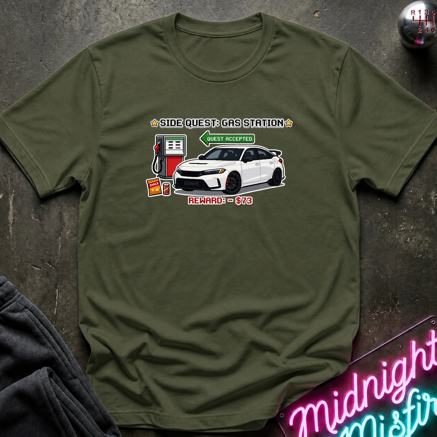 An olive green short-sleeve t-shirt with a graphic print of a white sports car and a gas pump, styled flat on a dark textured surface with a neon sign and dark fabric in the corners.