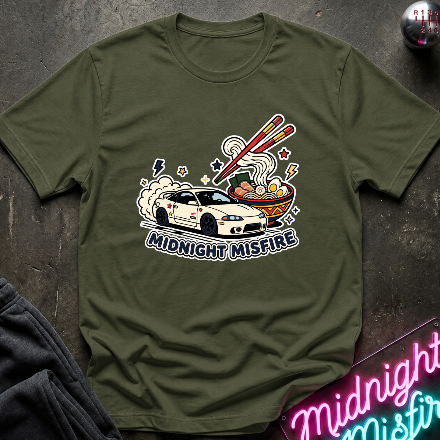 An olive green short-sleeve t-shirt with a colorful graphic of a white sports car racing toward a bowl of ramen with chopsticks, under the text 'MIDNIGHT MISFIRE'. The shirt is laid flat on a dark textured surface with a neon sign and dark pants partially visible.
