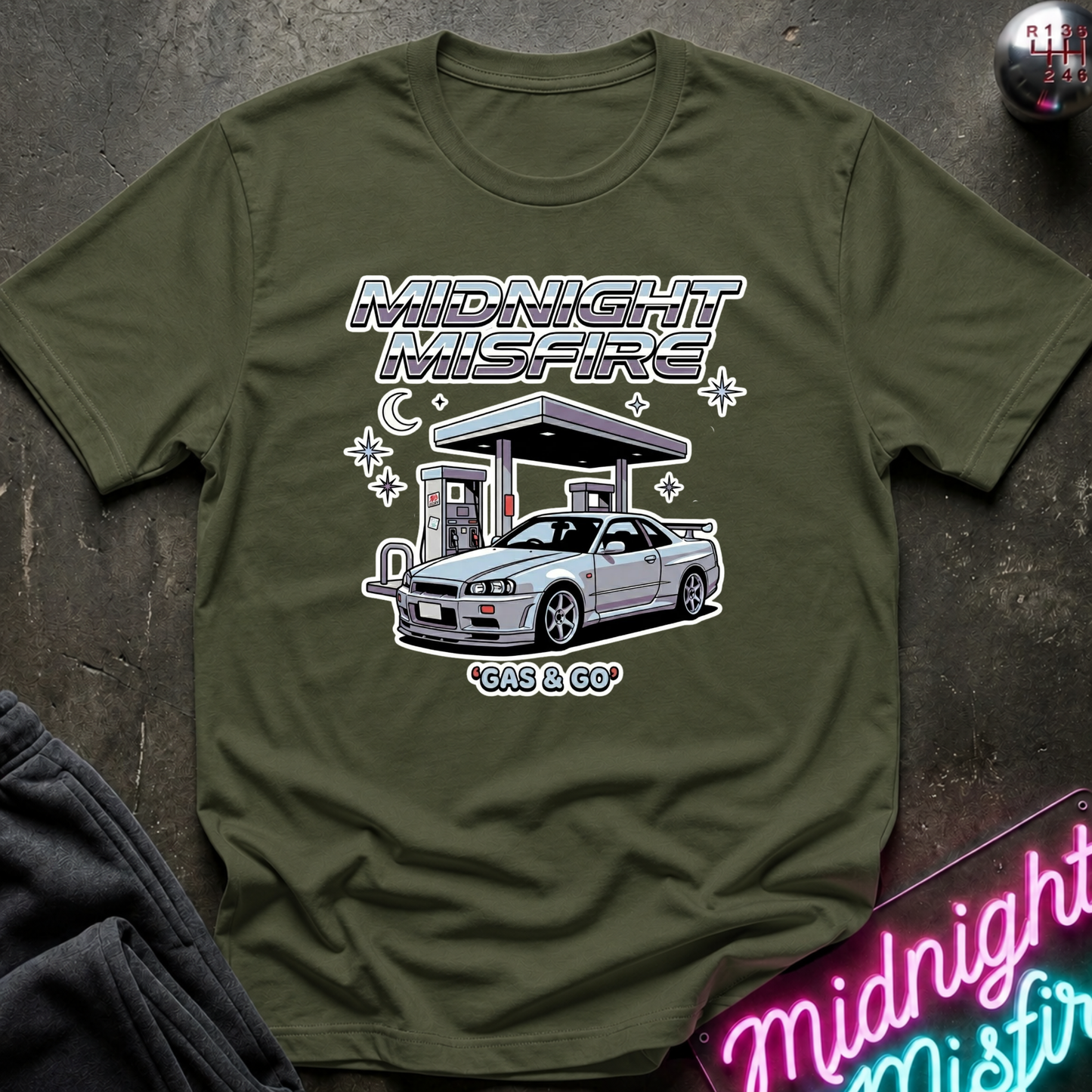 An olive green graphic t-shirt laid flat on a dark textured surface. The shirt features a white and silver graphic of a Nissan R34 at a gas station with the text 'MIDNIGHT MISFIRE' and 'GAS & GO'.