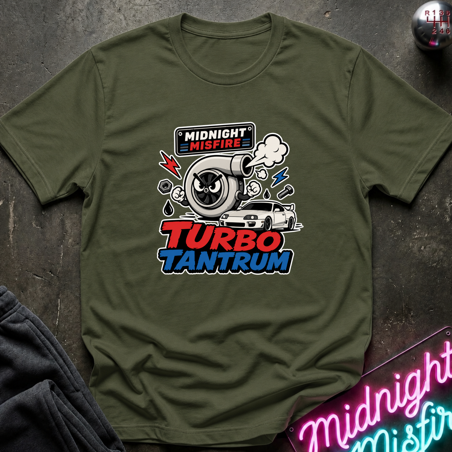 An olive green short-sleeve t-shirt with a 'Midnight Misfire Turbo Tantrum' graphic on the chest, displayed flat on a dark textured surface with a neon sign and dark sweatpants in the background.