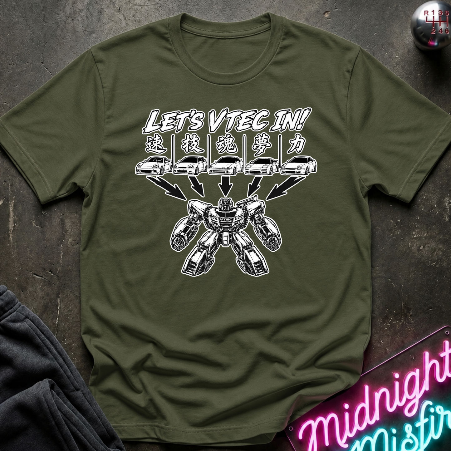 An olive green graphic t-shirt laid flat on a dark textured surface. The shirt features a white graphic of cars transforming into a robot with the text 'LET'S VTEC IN!'