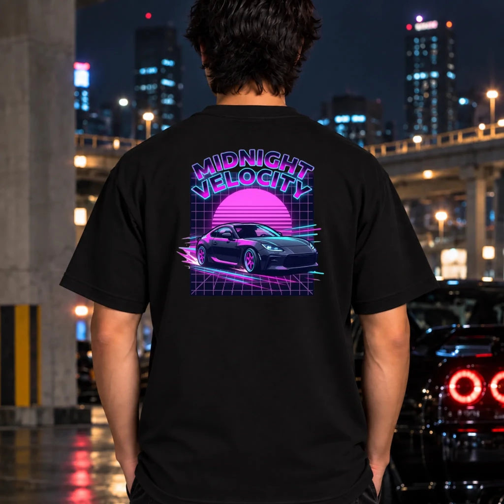 black short-sleeve t-shirt laid flat on a dark textured surface, featuring a colorful 80s retro-style graphic on the back with the text 'MIDNIGHT VELOCITY' and a pink neon car illustration
