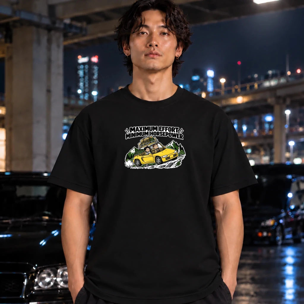 black graphic t-shirt laid flat on a dark textured surface. The graphic features a yellow sports car with a driver and passengers, with the text 'MAXIMUM EFFORT MINIMUM HORSEPOWER' above it.