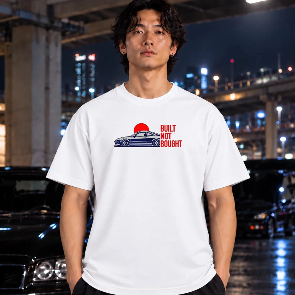 A man wearing a white graphic t-shirt with a blue car and red sun graphic, standing in front of a blurred night city background with cars.