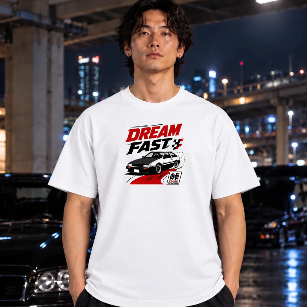A man wearing a white graphic t-shirt with a 'DREAM FAST+' racing design, standing in a city environment at night with blurred cars and lights in the background.