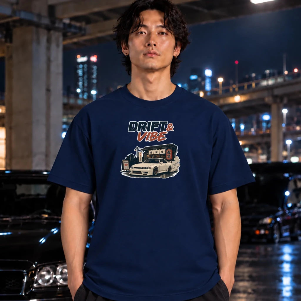 A man wearing a navy blue graphic t-shirt with a 'DRIFT & VIBE' car design, standing in a city parking lot at night.