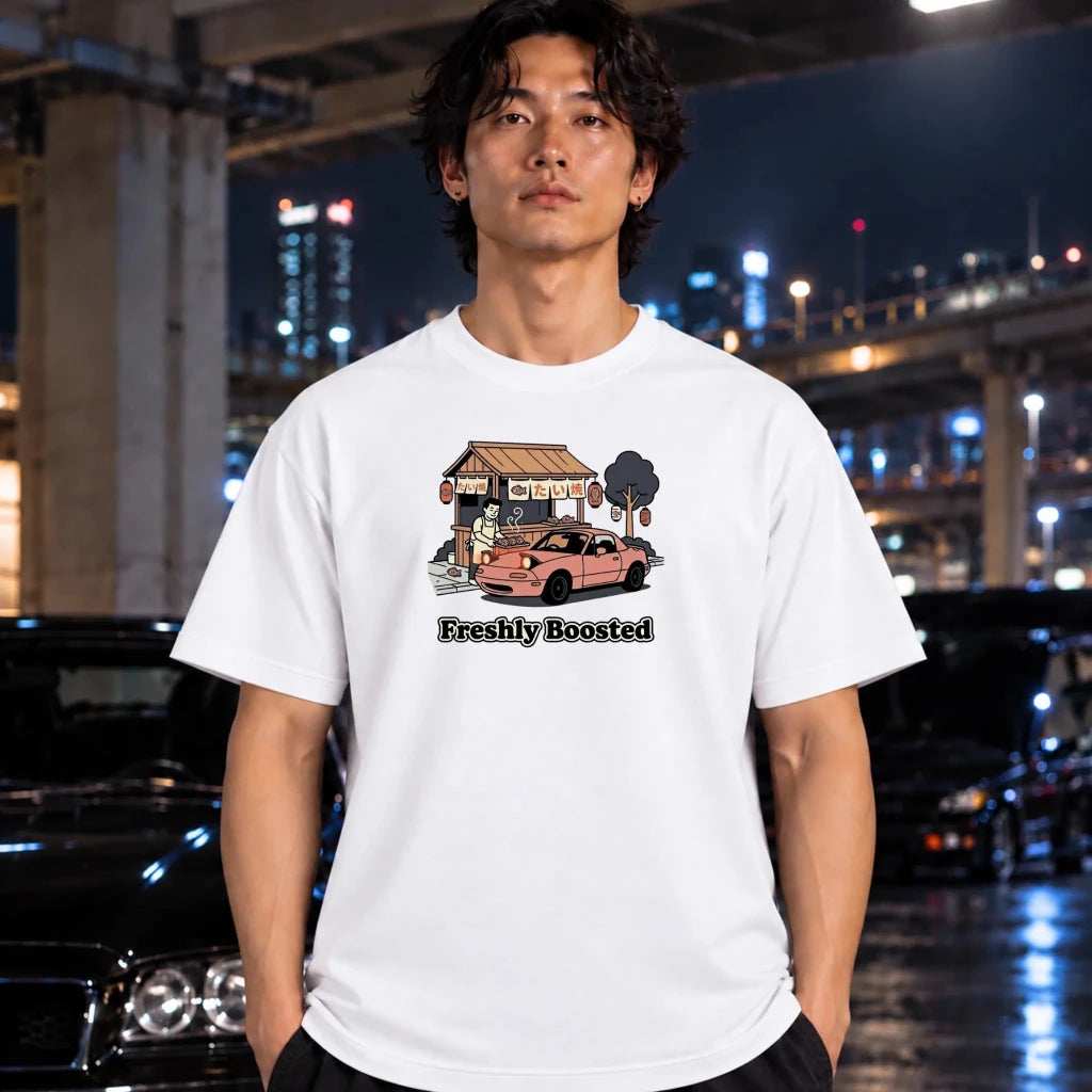 A man wearing a white graphic t-shirt with a cartoon illustration of a pink car and the text 'Freshly Boosted', standing in a nighttime urban setting with blurred cars and city lights in the background.