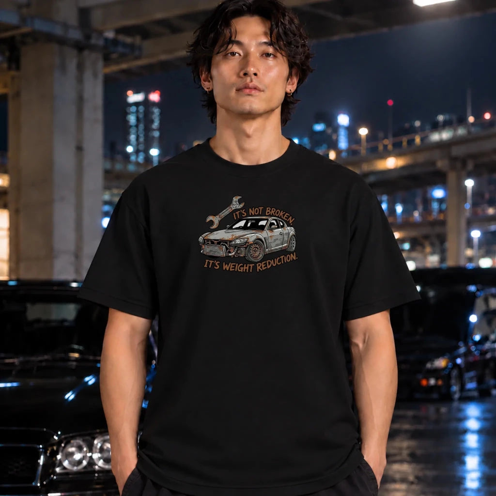 A man wearing a black graphic t-shirt with a cartoon car and the text 'IT'S NOT BROKEN IT'S WEIGHT REDUCTION.' standing in a nighttime urban setting with blurred cars and city lights.