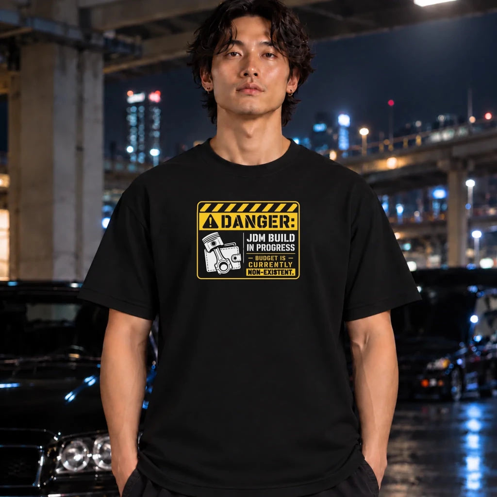A man wearing a black graphic t-shirt with a yellow and black 'DANGER: JDM BUILD IN PROGRESS' print, standing in a wet urban night scene with blurred cars and city lights in the background.