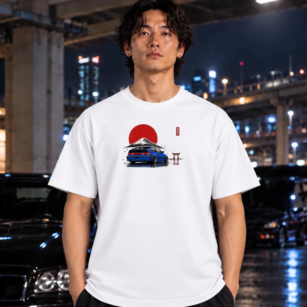 A man wearing a white graphic t-shirt with a blue car and red sun design, standing in a city environment at night.