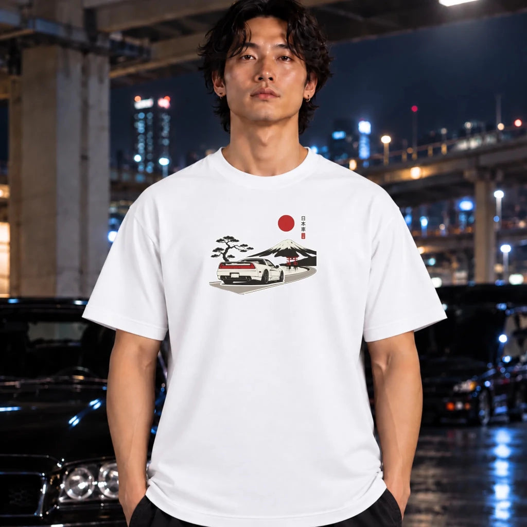A man wearing a white graphic t-shirt with a vintage Japanese car illustration, standing in front of a blurred night cityscape with cars.