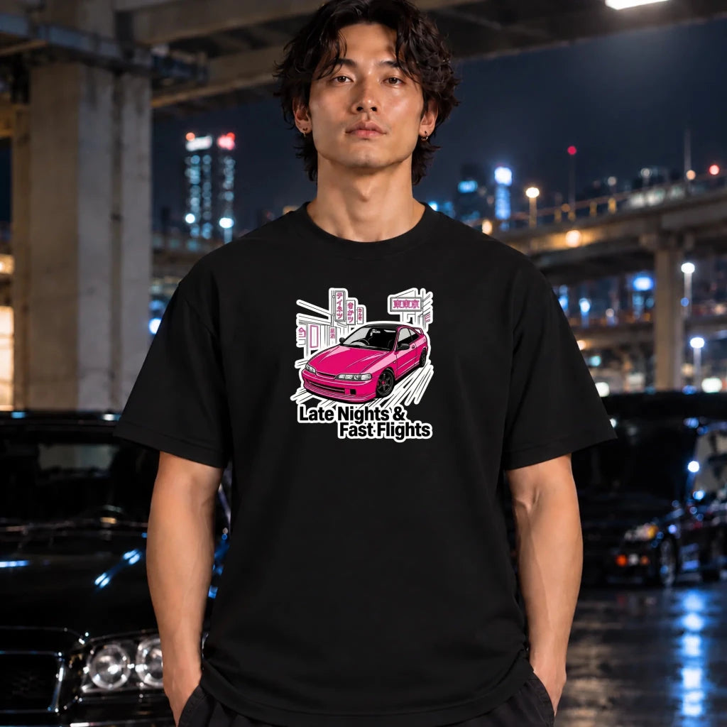 A man wearing a black graphic t-shirt with a pink car and the text 'Late Nights & Fast Flights' stands in a nighttime urban setting with blurred cars and city lights in the background.