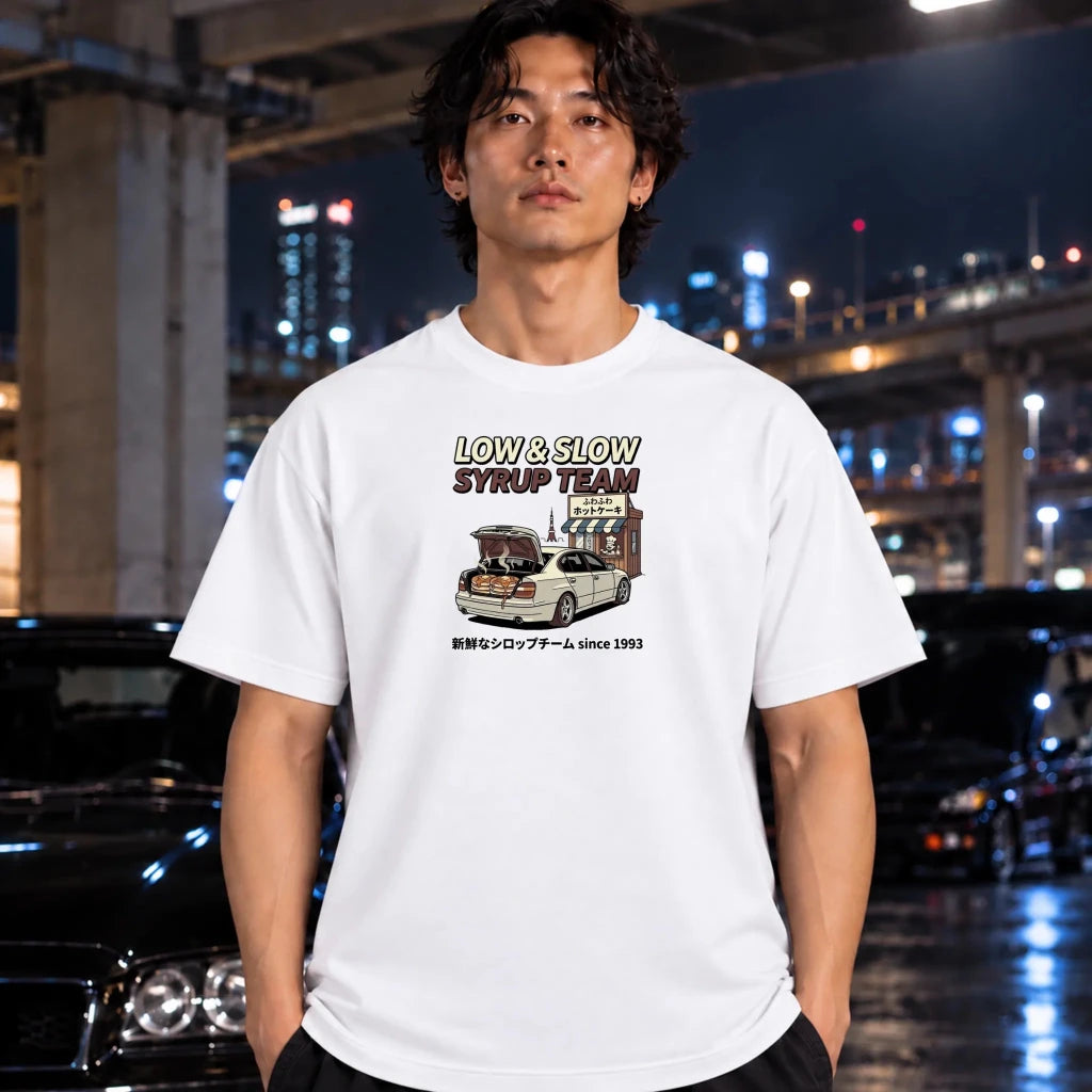 A man wearing a white graphic t-shirt with a 'LOW & SLOW SYRUP TEAM' car design, standing in a wet, city street at night with blurred cars and lights in the background.