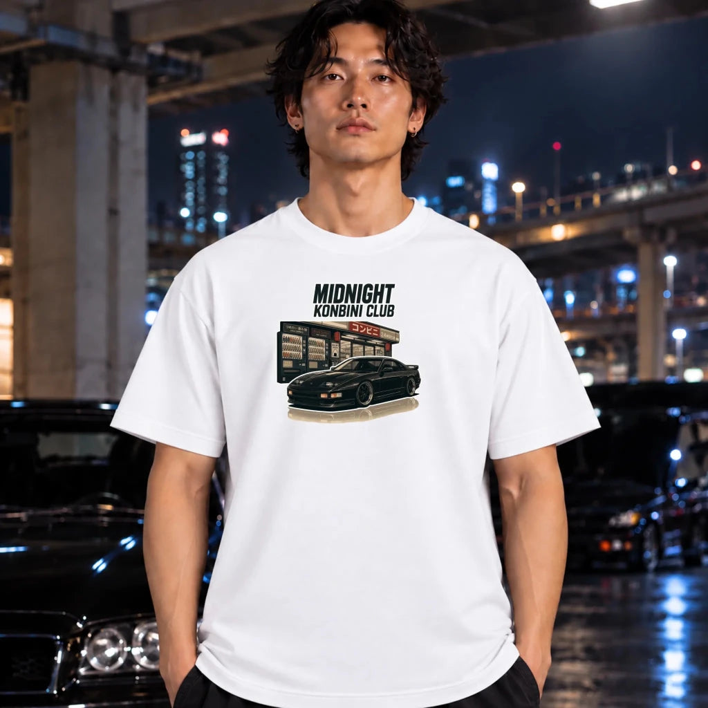 A man wearing a white graphic t-shirt with a 'Midnight Konbini Club' car print, standing in a wet urban night setting with blurred cars and lights in the background.