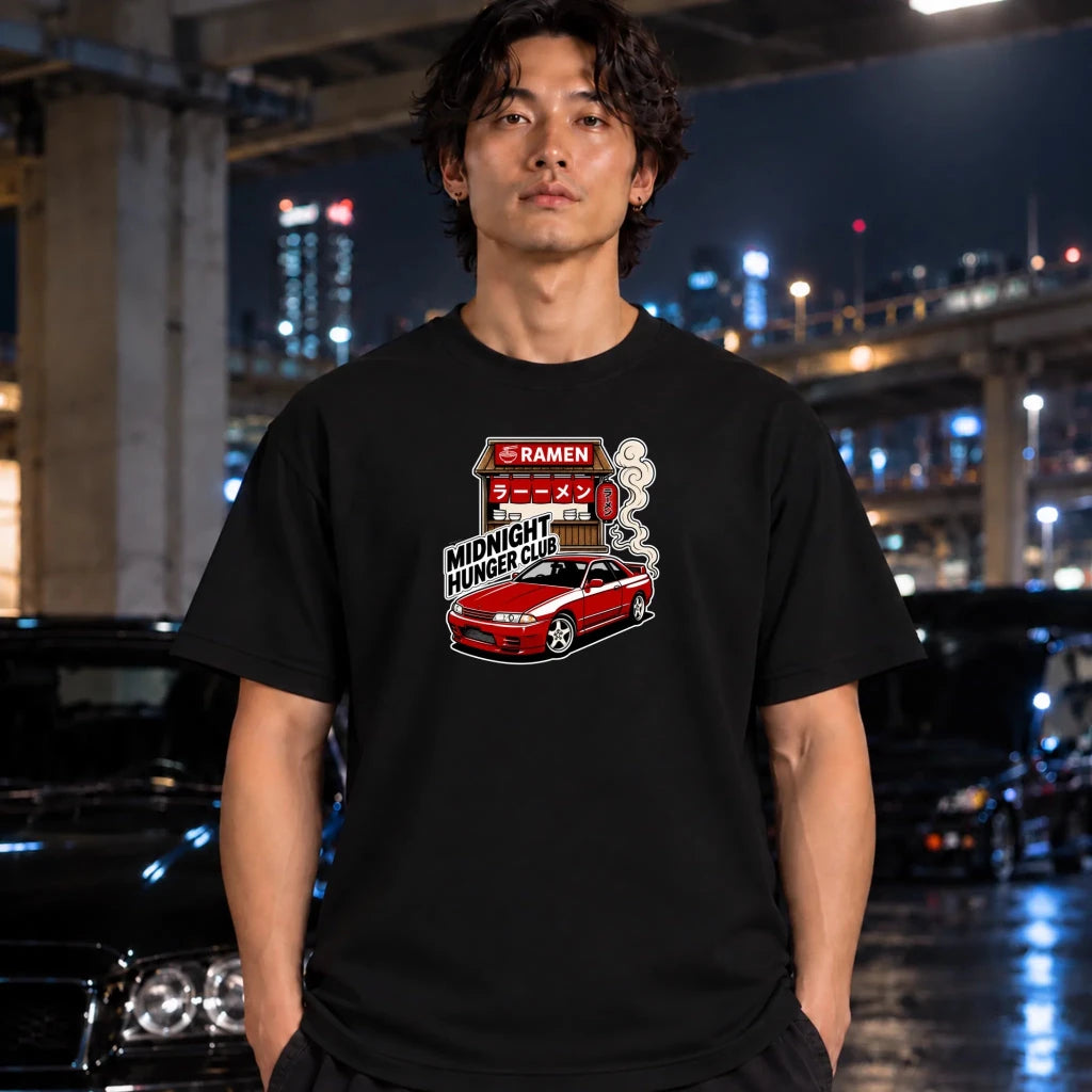 A man wearing a black t-shirt with a 'Midnight Hunger Club' ramen-themed graphic, standing in front of a blurred night city background with cars.