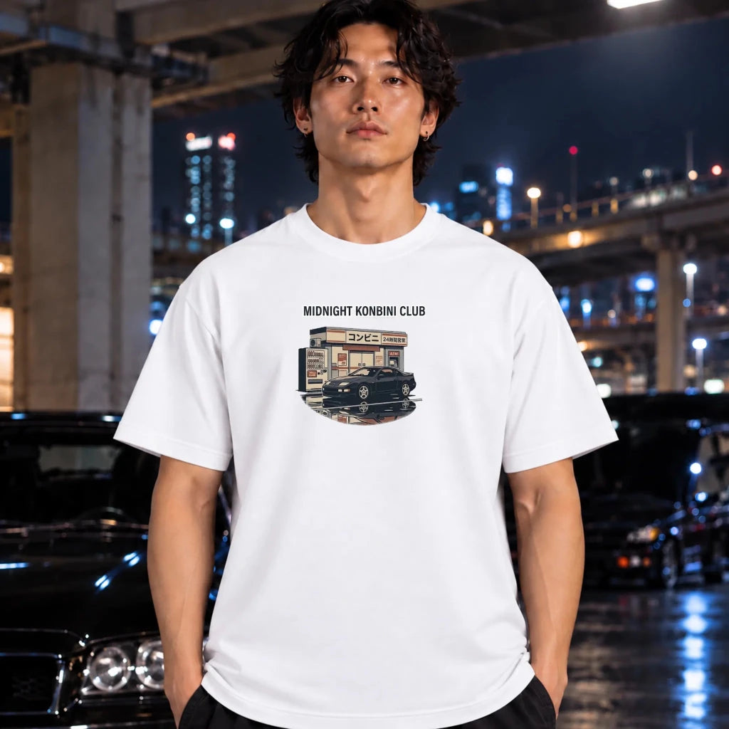 A man wearing a white graphic t-shirt with a 'Midnight Konbini Club' print, standing in front of a blurred night city background with cars.