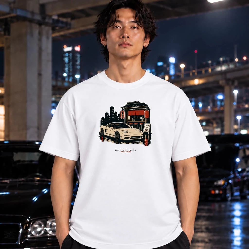 A man wearing a white graphic t-shirt with a vintage-style car illustration, standing in a city environment at night with blurred cars and lights in the background.