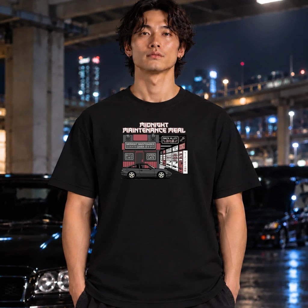 A man wearing a black graphic t-shirt with a 'Midnight Maintenance" print, standing in a wet, nighttime urban environment with blurred cars and city lights in the background.