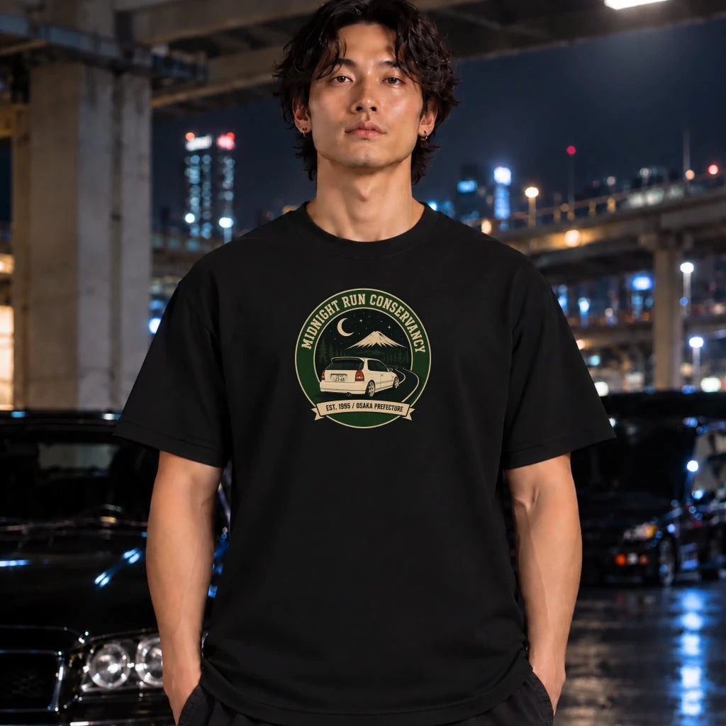 A man wearing a black crewneck t-shirt with a circular green and beige 'Midnight Run Conservancy 1995' graphic, standing in a nighttime urban setting with blurred cars and city lights in the background.