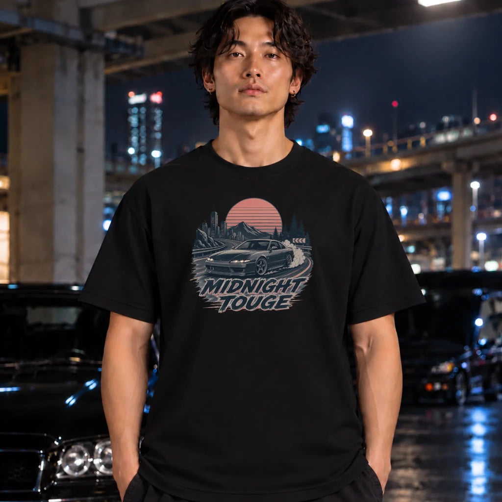 A man wearing a black graphic t-shirt with a retro-style 'Midnight Touge' car design, standing in a wet urban night environment with blurred cars and city lights in the background.