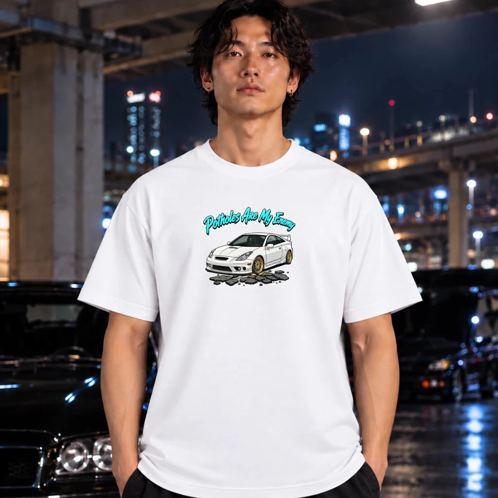 A man wearing a white graphic t-shirt with a printed design of a white car and the text 'Potholes are my enemy' stands in front of a blurred night city background with cars.