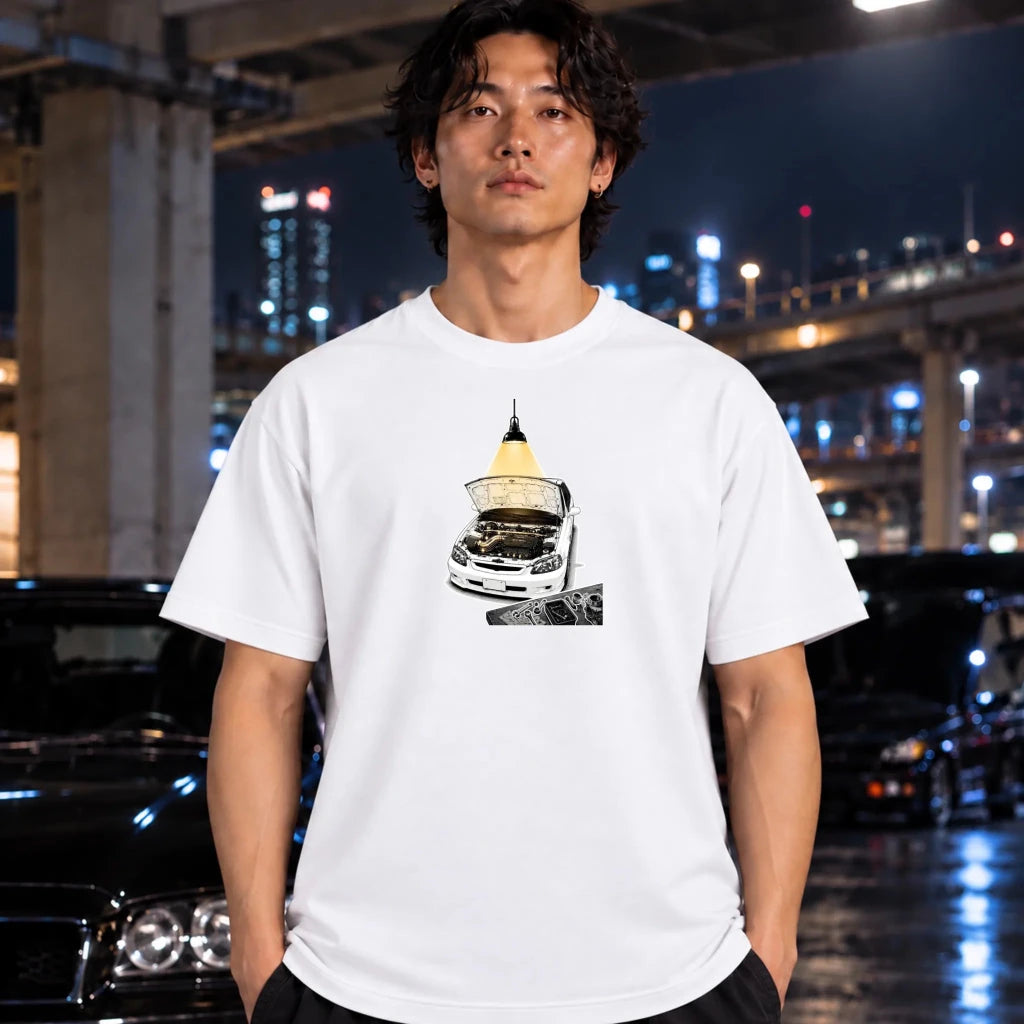 A man wearing a white graphic t-shirt with a car and engine design, standing in front of a blurred night city background with cars.