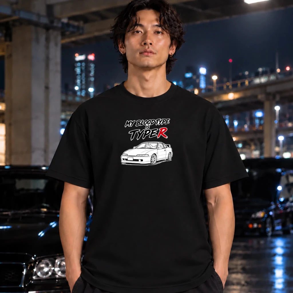 A man wearing a black graphic t-shirt with a white and red car design and the text 'MY BLOOD TYPE IS TYPE R' stands in front of a blurred night city background with cars.