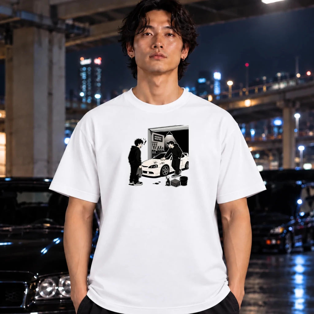 A man wearing a white short-sleeve graphic t-shirt with a black and white illustration of a person standing next to a white sports car. The background is a blurred night scene of a city street with lights and concrete pillars.